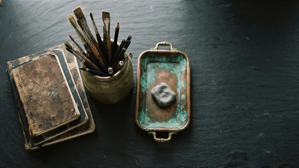 Worn sketchbooks stacked beside a jar of used brushes and pencils and a patinated copper tray holding a kneaded eraser on a dark slate table