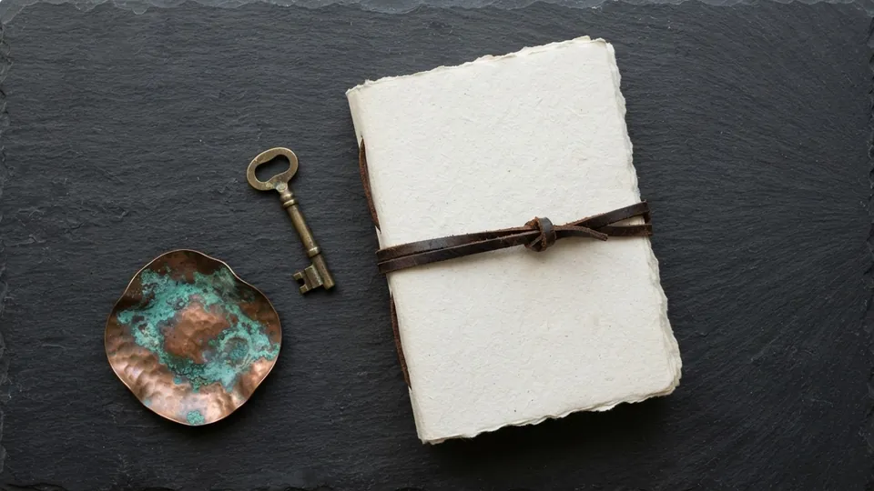 Closed sketchbook tied shut with a leather cord, an old brass key and a verdigris copper dish on dark slate