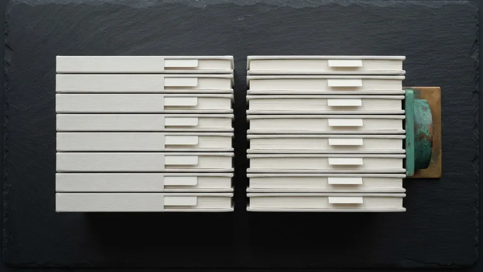 Two neat stacks of sketchbooks with plain paper tabs held by a patinated copper bookend on dark slate, an archive of tutorials