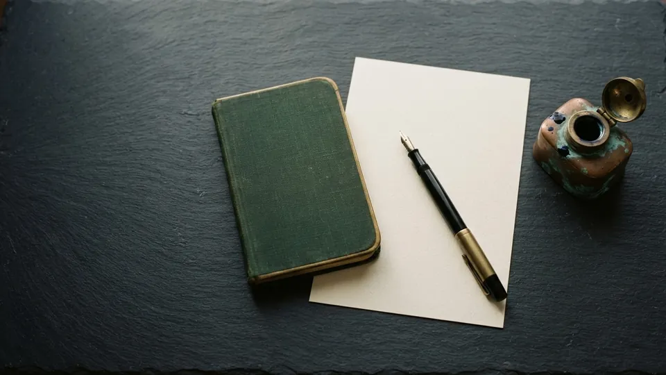 Blank sheet of writing paper with a fountain pen across it beside a green cloth notebook and a verdigris copper inkwell on dark slate